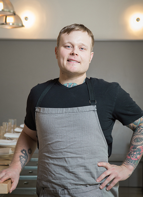 A photo of Chef Brian Clevenger with one hand on his hip and the other hand resting on a counter wearing a grey apron.