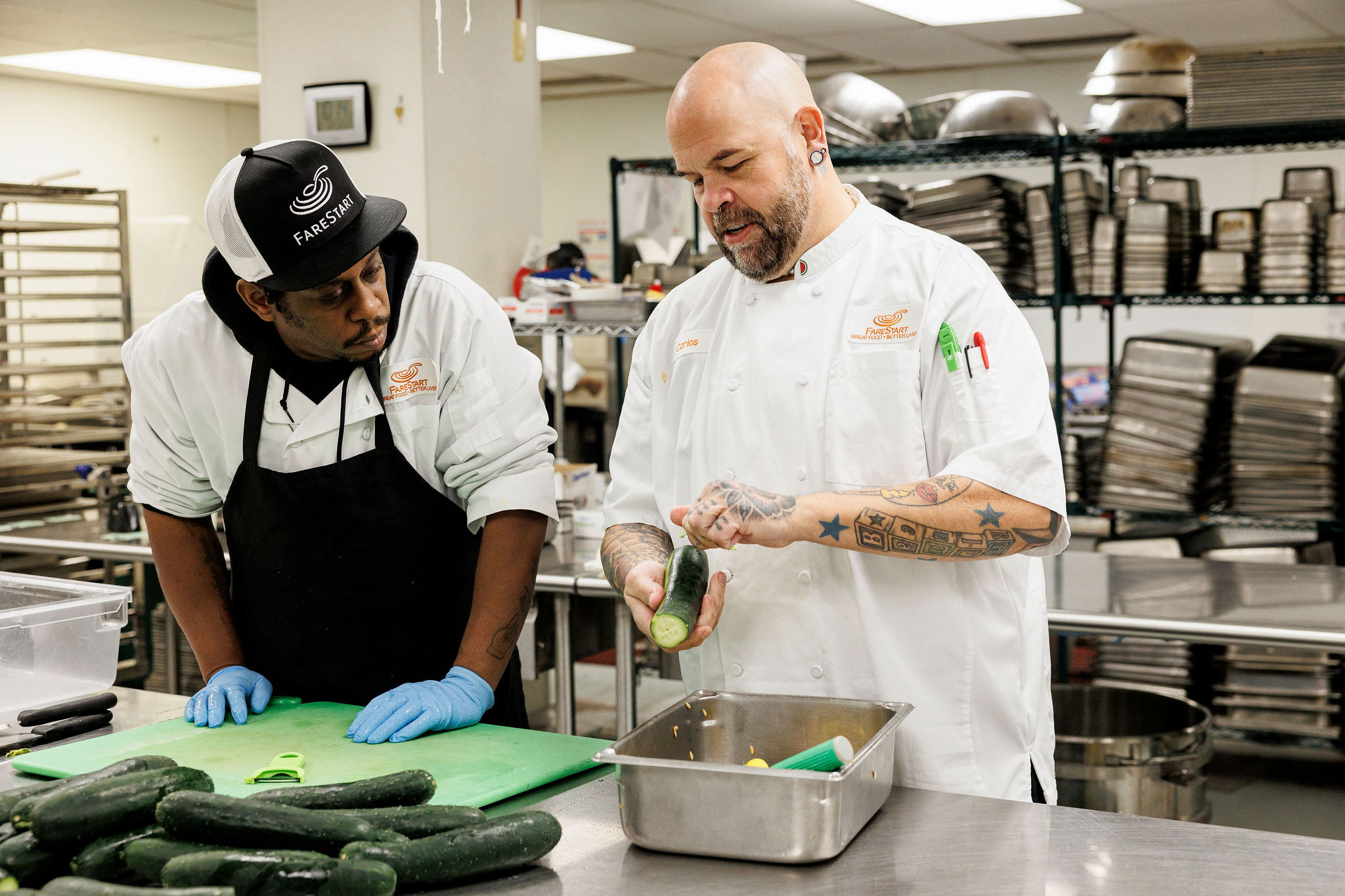 A student looks on as Chef Carlos, a trainer at FareStart, demonstrates a technique on a cucumber in a commercial kitchen setting