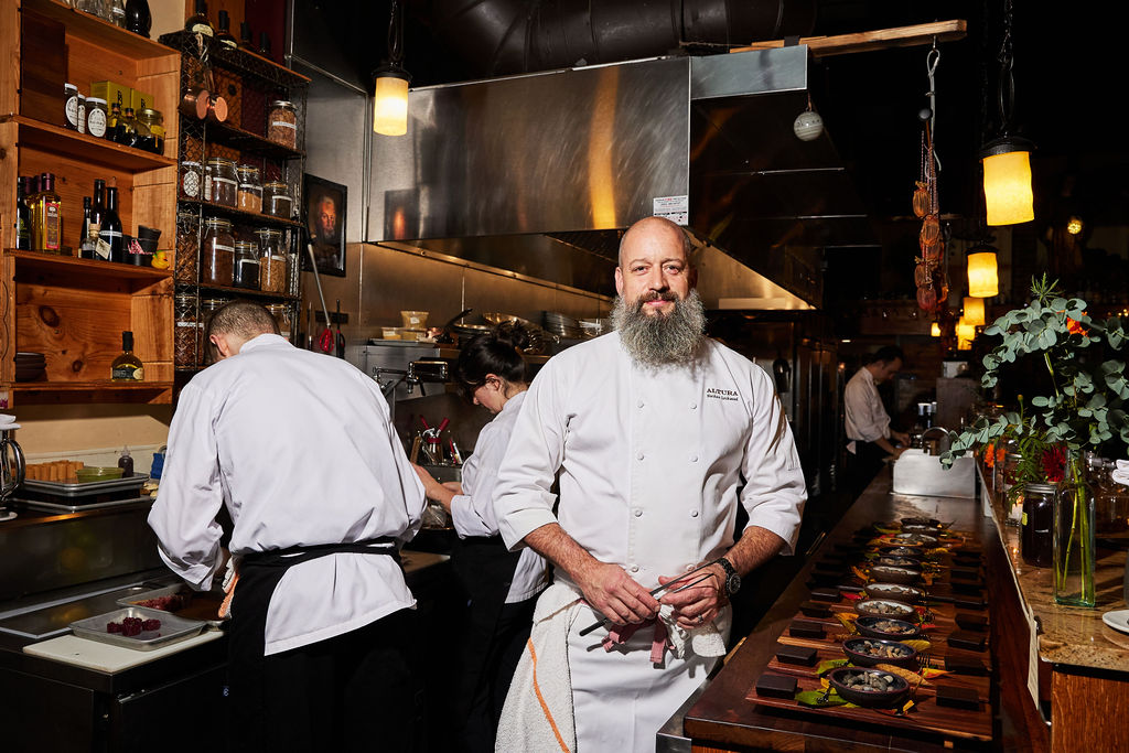 Chef Nathan Lockwood standing in a kitchen smiling next to dishes he is preparing to serve