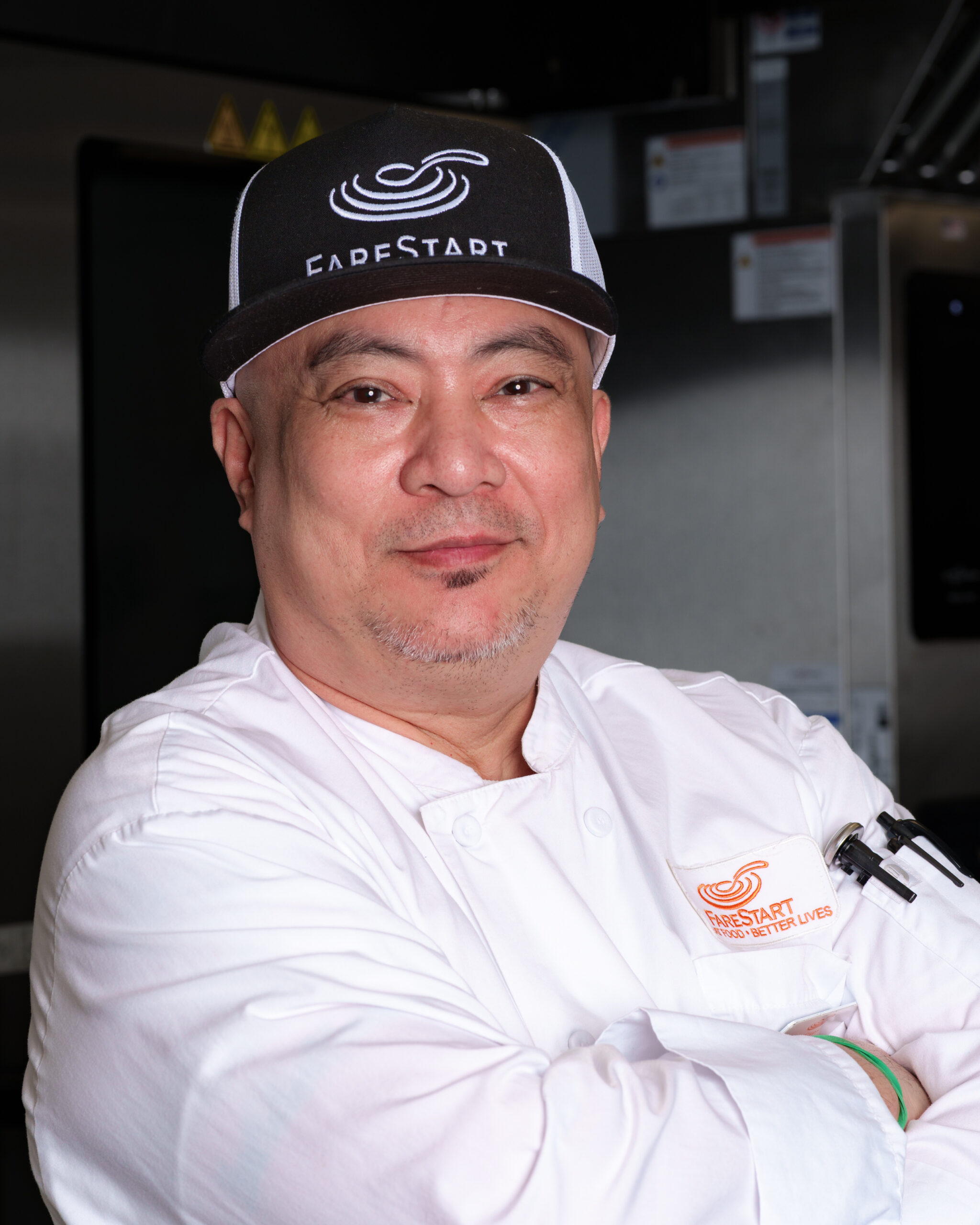 Anthony (he/him) Food Pathways Program Graduate standing in the kitchen with his arms crossed wearing a FareStart hat and chef